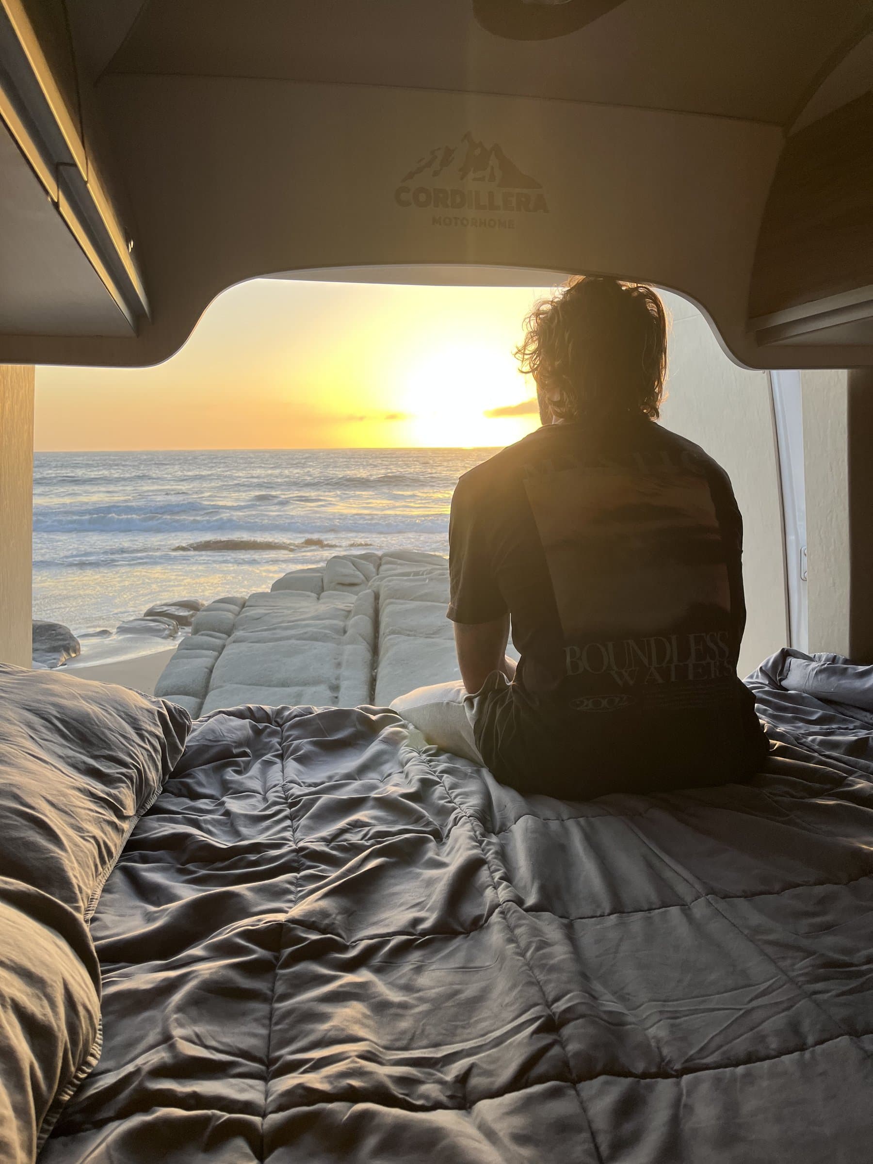 Vista del atardecer al mar desde el interior del motorhome con una persona sentada en la cama